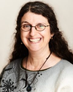 A white female with long wavy hair. She is wearing a grey embroidered top and a necklace.